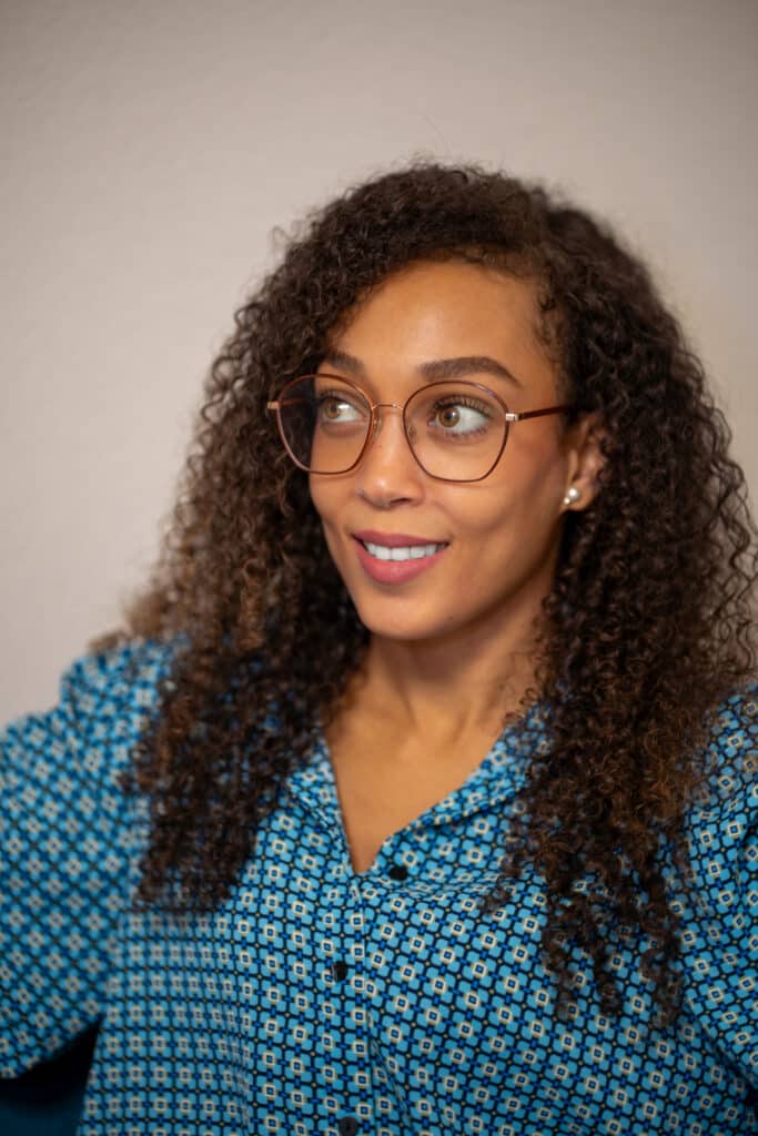 Lächelnde Frau mit lockigem Haar, Brille, blauem Hemd.